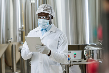 African American specialist in white coat and mask using tablet pc during his work at factory