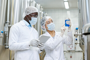 Multiethnic couple of scientists conducting research in team at factory, they examining blue liquid...