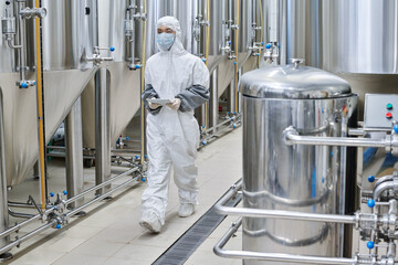 Worker in protective wear using digital tablet while walking along the workshop at chemical factory