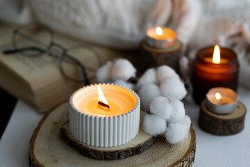 Cozy autumn or winter composition with aromatic candle, wool sweater, cotton, book. Aromatherapy, home atmosphere of cosiness and relax. Wooden background close up.