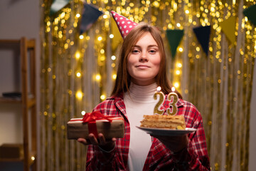 Young woman holding plate with tasty birthday cake and gift at party background. 23th birthday