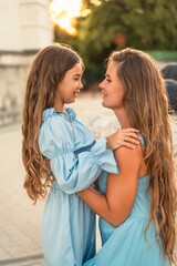 Fototapeta premium Portrait of a mother and daughter in blue dresses with flowing long hair against the backdrop of a sunset and a white building. They look at each other. Family stories on the weekend