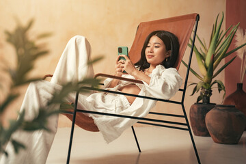 Korean girl in stylish boho chair using phone, sitting in minimalistic rustic decor on resort