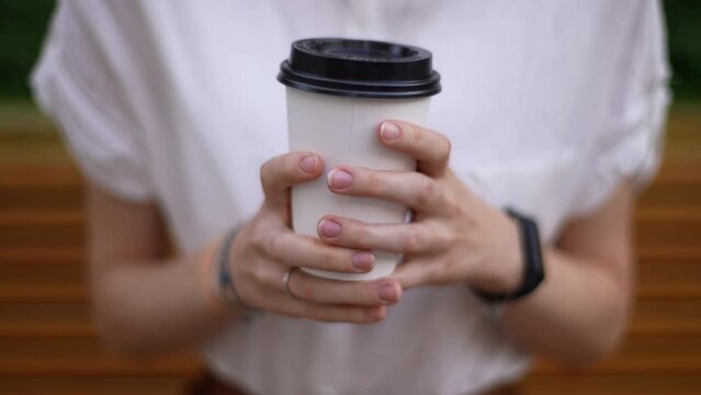 Close-up cropped shot of unrecognizable young woman holding in hands takeaway cup of hot coffee near chest while waiting someone in city street in summer day. Shooting in slow motion.