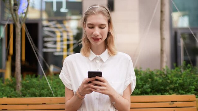 Portrait of happy positive young beautiful woman using smartphone sitting on bench outside then smiling and looking at camera. Front view of cheerful lady using mobile phone relaxing on city street.