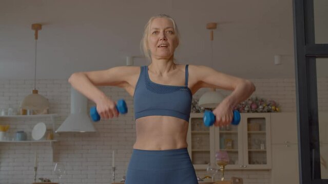 Determined motivated beautiful sporty fit mature female exercising dumbbell upright row, strengthening deltoids, trapezius, and triceps while practicing indoor sports training.
