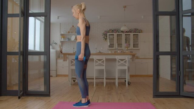 Determined charming fitness senior woman in activewear practicing cross training, performing reverse lunge with dumbbells exercise, strengthening core, glutes, and hamstrings in domestic room.