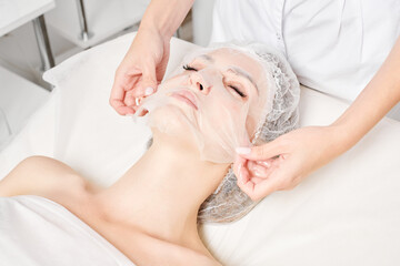 Beautician smoothes sheet mask on woman face for rehydrate face skin, anti wrinkles cosmetic...