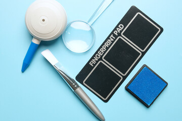 Flatlay picture of toy fingerprint pad, magnifying glass, puffer, brush and inkpad on blue background.