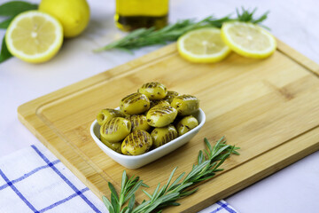 Grilled green olives with rosemary, olive oil, lemon and spices in a bowl on a wooden cutting board on the table.
Copy space. Close up. Mediterranean food. Turkish olives. Grilled olives in a plate.