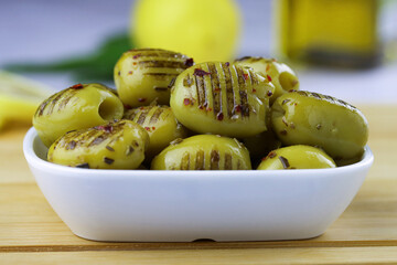 Grilled green olives with olive oil, lemon and spices in a bowl on a wooden cutting board on the table.
Close up. Mediterranean food. Turkish olives. Grilled olives in a plate. Selective focus