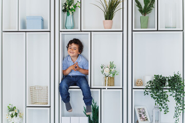Fototapeta na wymiar Naughty curly hispanic boy in blue jeans, polo, sitting on shelve among vases with flowers and plants looks at camera, smiles. Adorable caucasian kid posing at home. Childhood, eco friendly concept.