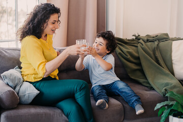 Happy caucasian curly boy smiles sitting with mom on couch having fun, clinks glass of water with...