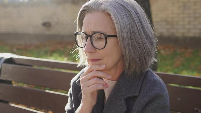 Nervous gray-haired lady in elegant coat and eyeglasses sitting on bench in park and waiting for boyfriend she met on dating web site in internet, blind date, fear to be alone