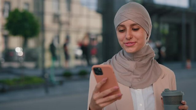 Happy arabian businesswoman wearing hijab walking near the office centre on a break using smartphone, texting with friends holding the cup of coffee. People and technology business concept