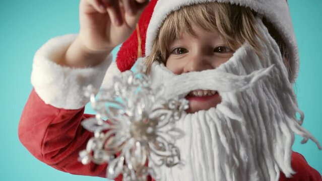 Lovely little child in Santa Claus costume, artificial beard playing with snowflake Christmas tree toy. Festive mood. Happy childhood, kids, funny son