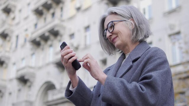 Attractive senior woman in fashionable coat adjusting her hairstyle, chatting on smartphone with boyfriend, flirting, desire to be beautiful and happy