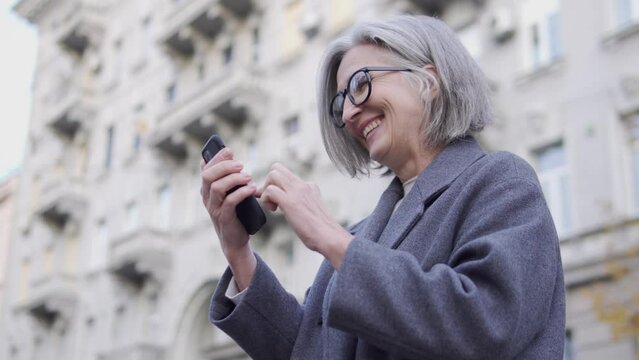 Senior woman in elegant eyeglasses and coat laughing reading messages on smartphone, waiting for meeting with friends outdoor, using free wi-fi connection