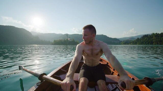 Young man with bare torso sits in wooden boat sailing to small island with catholic church on Bled lake against high mountains of Slovenia