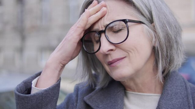 Exhausted mature woman in big eyeglasses touching forehead, feeling dizziness and pain in head, general weakness and fatigue of the body, first symptoms of cold or flu, emotional burnout