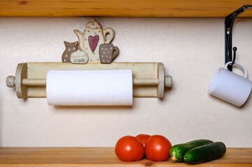On the kitchen wooden shelf are several red tomatoes and two large green cucumbers. A wooden fixture with a paper towel is installed on the wall. A white mug is hanging on the hook.