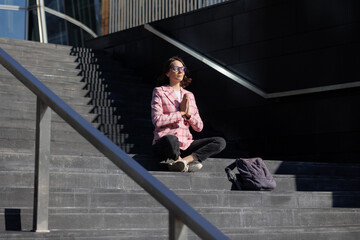 business woman in pink jacket sits on the steps of office in lotus position and meditates, entrepreneur relieves stress and tension. startup female is successful confident, interviewing negotiating