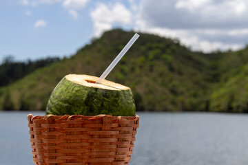 Green coconut ready to drink with a straw on a sunny day in a wicker basket with blurred lake and mountains  in the background