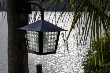 Small lamp attached to a wooden pole among the vegetation with blurred lake illuminated by the sun in the background