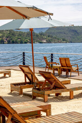 Several empty wooden lounge chairs on the sand under green umbrellas. In the background, a huge lake. Vertical