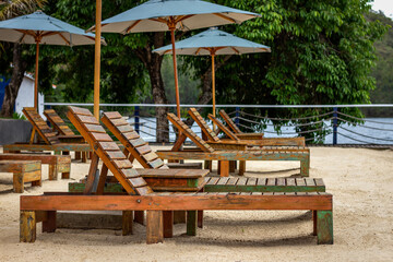 Several empty wooden lounge chairs on the sand under green umbrellas. In the background, a large tree and lake water
