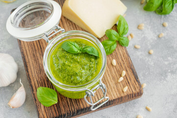 Traditional Italian basil pesto sauce in a glass jar on a board with ingredients for cooking on a gray concrete background. Copy space.