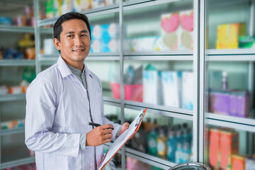 Asian pharmacist in uniform holding pen and clipboard standing in front of storefront