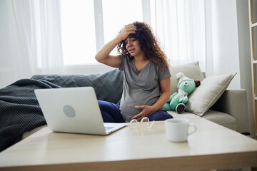 A pregnant woman with a headache sits at home on the sofa with a laptop and watches TV shows movies and series. Lifestyle of a pregnant woman, preparation for childbirth, last month of pregnancy