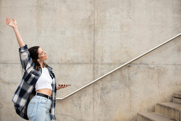 young caucasian girl with straight black hair, casual black and white plaid shirt and jeans, dancing alone in the street with headphones and cell phone. arm up.