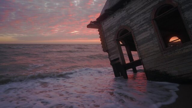 Mysterious vintage flooded leaning wooden house washed by sea waves, at coastline on spectacular pink sunset. Enigmatic rickety building sinking in water on sandy beach on dreamy cinematic sunrise.