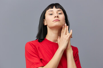 a sweet, gentle, sophisticated, relaxed woman with black hair stands in a red T-shirt on a gray background and holds her hands near her face