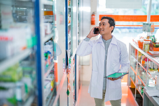 male pharmacist in uniform making phone call while holding pad in pharmacy