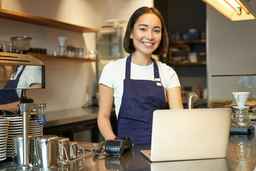 Portrait of young girl cafe owner, looking at her laptop, taking order, serving customer in coffee...