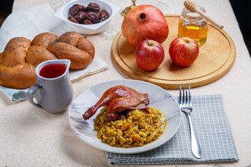 Rosh Hashanah next to pomegranate honey apples wine and challah on a white table and candles next to a duck's leg and spelt in a plate.