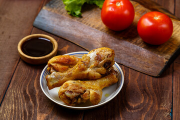 Baked chicken drumsticks on a plate on a wooden table next to soy sauce, herbs and tomatoes.