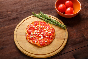 Homemade pizza with tomatoes and mozzarella on a round wooden board next to a sprig of rosemary and tomatoes.