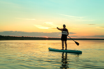 A man in shorts on a SUP board with a paddle at sunset swims in the water of the lake against the backdrop of the sunset sky.
