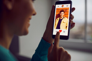 Online dating. Smiling woman clicks like button on handsome young man in social network or online dating app. Close up of mobile phone in hands of woman pressing red heart button with her finger.
