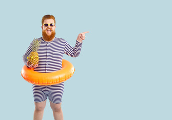 Young redhead happy Caucasian funny fat man tourist with life buoy on belt smiling pointing finger to side holding pineapple dressed in striped beach suit stands. Summer resort concept - Powered by Adobe