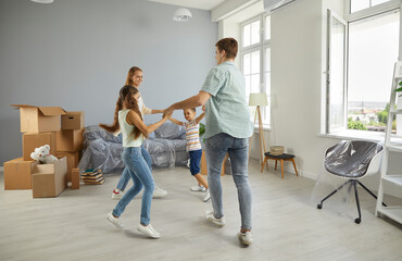 Cheerful young family with children dancing and having fun while moving to new apartment. Mom, dad, son and daughter are holding hands in circle near cardboard boxes and covered furniture.