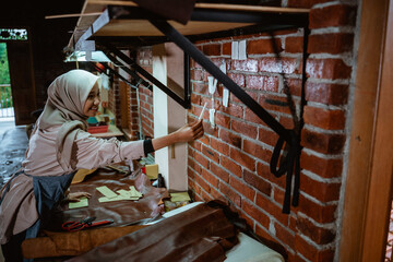 Veiled female leather worker making small notes on the wall while standing in her studio