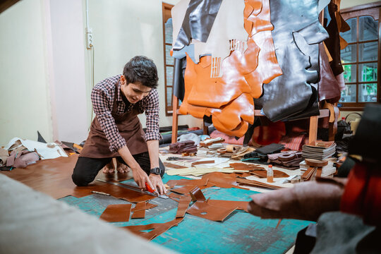 Craftsman wearing apron cutting leather with knife on wooden floor in workshop