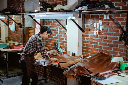 Asian craftsman in apron working with leather in workshop, leather craft making process