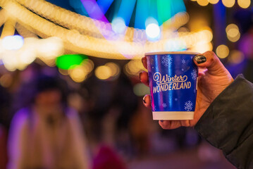 A hand holding a winter wonderland cup with blurred background of the Hyde Park Winter wonderland festival in London