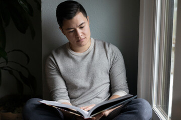 Fototapeta premium Asian American trans man sits by the windowsill.